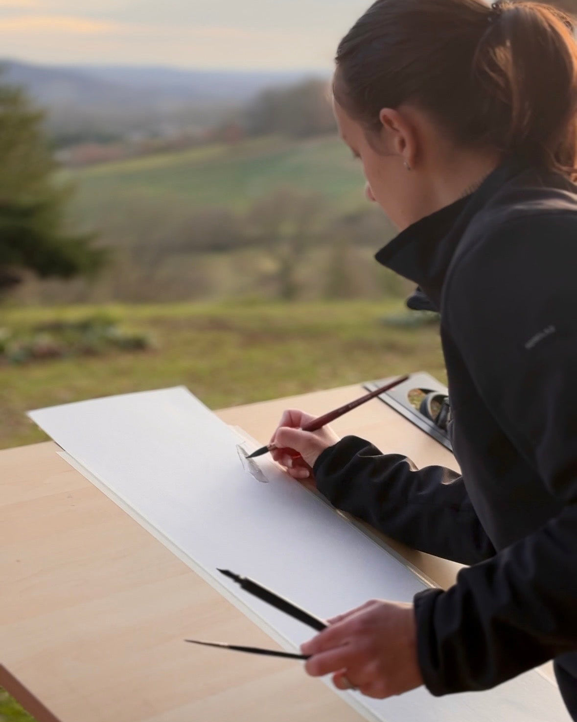 Sur cette image, on peut voir l'artiste Coline Pratviel en train de commencer une peinture sur sa table à dessin à l'extérieur, derrière elle se trouve la vallée où le soleil commence à se coucher dans une ambiance douce et inspirante