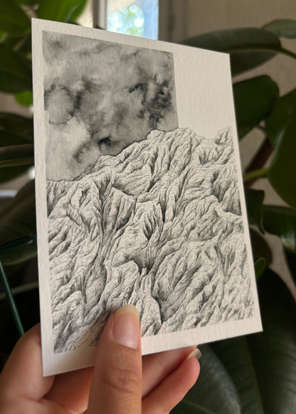 Vue de profil du Tirage fine art de la montagne contrôle réalisée à l'encre de chine en noir et blanc au format carte postale 10 x 15 cm tenu par l'artiste devant une plante verte