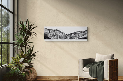 Reproduction de la peinture du massif du néouvielle dans les Pyrénées à l'encre de chine en noir et blanc sur une plaque de 33x95 d aluminium dibond pour un rendu moderne, mettant en valeur les profondeurs des nuances de gris, de blanc et de noir de ce tableau. Le tableau est affiché dans un salon lumineux à côté d'un fauteuil chaleureux et de plantes, ajoutant une touche d'élégance à cette décoration confortable, naturelle et chaleureuse.