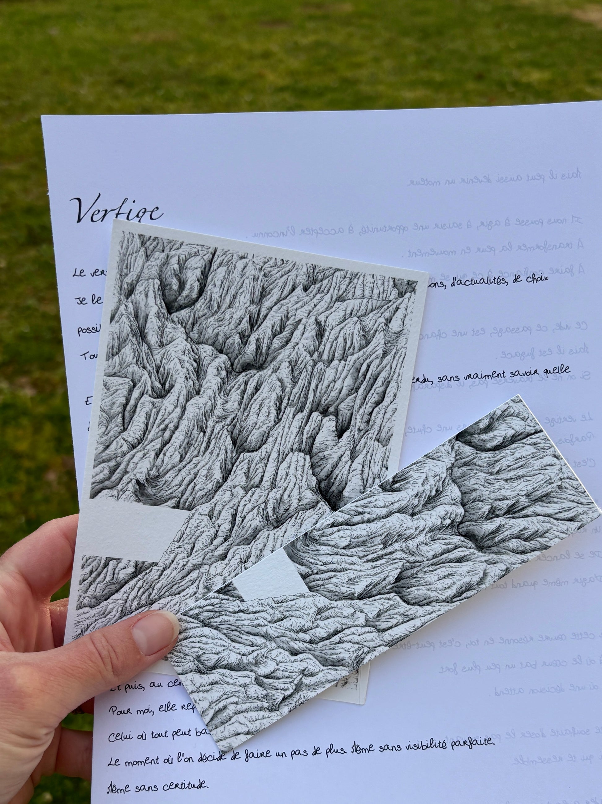 Apercu de la correspondance à l'encre des cimes du mois d'avril nommée vertige , ont peut voir sur cette image la lettre manuscrite du mois, l'impression fine art au format 10 x 15 cm ainsi que le marque page tiré de la peinture originale. Les 3 sont tenus par une main au-dessus d'herbe. idée cadeau montagne, zen, cadeau peinture noir et blanc, correspondance mensuelle physique 