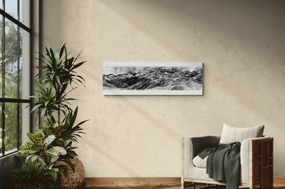 Reproduction de la peinture de la vue depuis le Pic de Bacanère  dans les Pyrénées à l'encre de chine en noir et blanc sur une plaque de 33x95 d aluminium dibond pour un rendu moderne, mettant en valeur les profondeurs des nuances de gris, de blanc et de noir de ce tableau. Le tableau est affiché dans un salon lumineux à côté d'un fauteuil chaleureux et de plantes, ajoutant une touche d'élégance à cette décoration confortable, naturelle et chaleureuse.