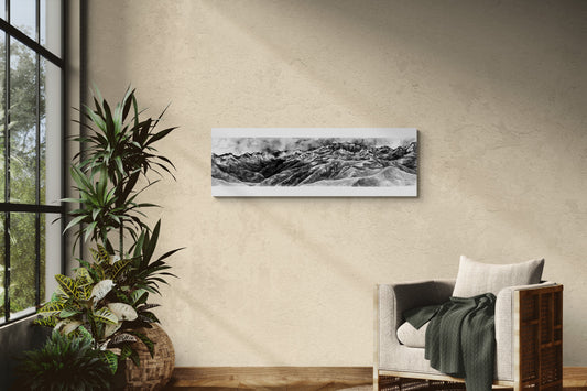 Reproduction de la peinture de la vue depuis le Pic de Bacanère  dans les Pyrénées à l'encre de chine en noir et blanc sur une plaque de 33x95 d aluminium dibond pour un rendu moderne, mettant en valeur les profondeurs des nuances de gris, de blanc et de noir de ce tableau. Le tableau est affiché dans un salon lumineux à côté d'un fauteuil chaleureux et de plantes, ajoutant une touche d'élégance à cette décoration confortable, naturelle et chaleureuse.