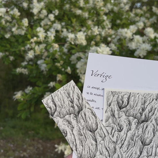 Vidéo de l'apercu de la correspondance à l'encre des cimes du mois d'avril nommée vertige , ont peut voir sur cette image la lettre manuscrite du mois, l'impression fine art au format 10 x 15 cm ainsi que le marque page tiré de la peinture originale. Les 3 sont tenus par une main au-dessus d'herbe et devant un buisson fleurit blanc, renforçant l'ambiance zen et naturelle de correspondance faite pour ralentir. idée cadeau montagne, correspondance mensuelle physique, encre de chine